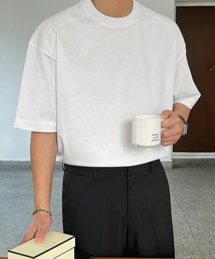 ホワイトの半袖Tシャツにブラックのスラックスを合わせ、白のマグカップを持って立つ男性の正面アップ。