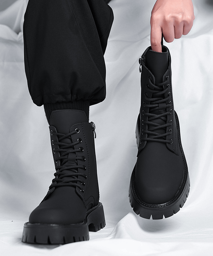 黒いパンツを穿いた人物が、片方のブラック厚底ブーツを着用し、もう片方のブーツをプルストラップを持って持ち上げている、着用時のサイズ感やシャフトの長さが伝わるフロントビュー写真