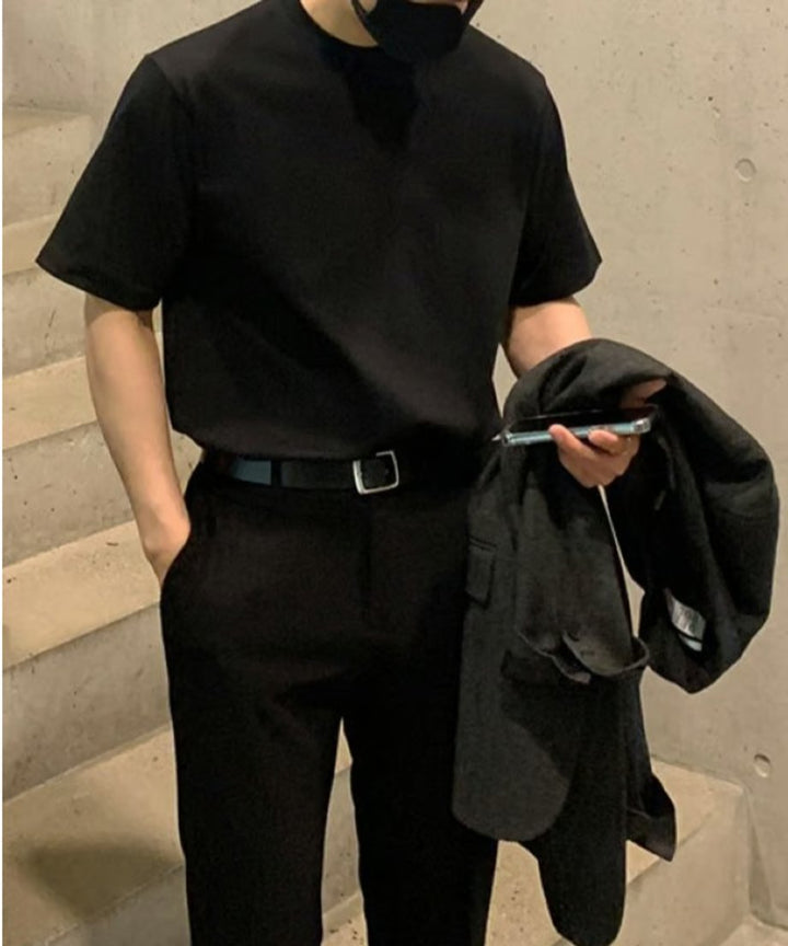 ブラックのクルーネックTシャツをブラックのパンツにタックインし、上着とスマートフォンを片手に持って階段に佇む男性の正面。