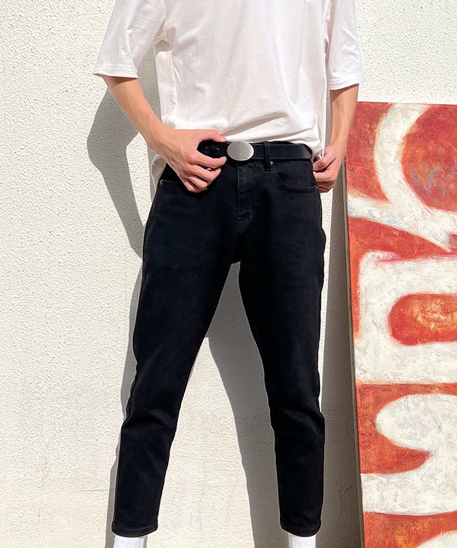 白い壁の前に立ち、白い半袖Tシャツと黒いスキニーパンツにシルバーオーバルバックルベルトを合わせた人物の全身スタイリングカット。