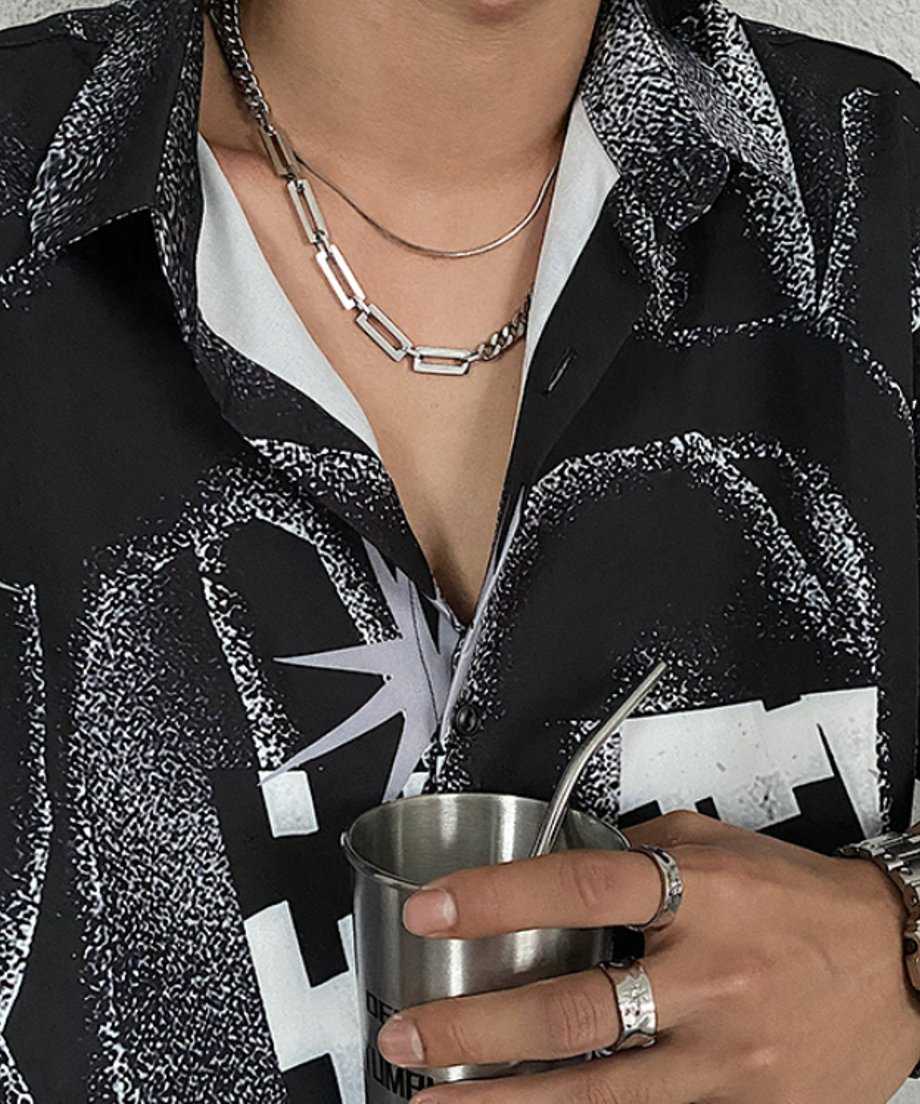 総柄シャツを着用し、メタルタンブラーを手にした男性の胸元で、細身のスネークチェーンとレイヤードされたコンビチェーンネックレスのスタイリングカット