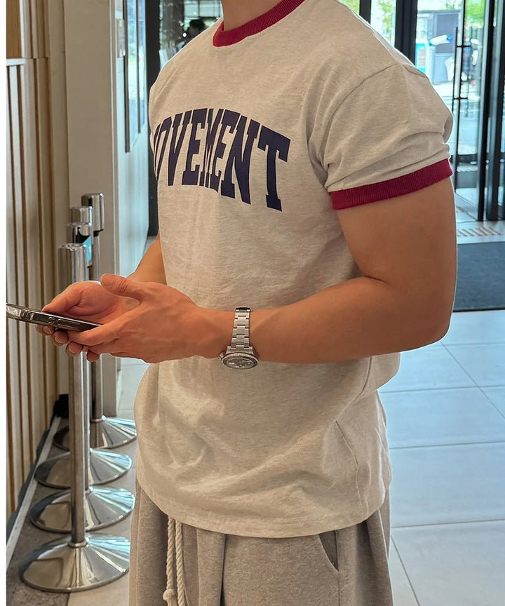 オートミール地のリンガーTシャツを着用し、メタルバンドの腕時計を付けた左手でスマートフォンを操作する男性の左側面からのアップカット。
