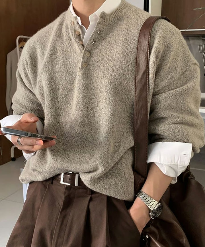 ベージュのヘンリーネックニットのポケットに片手を入れ、もう片方の手でスマートフォンを操作している男性の手元・胸元のアップカット。