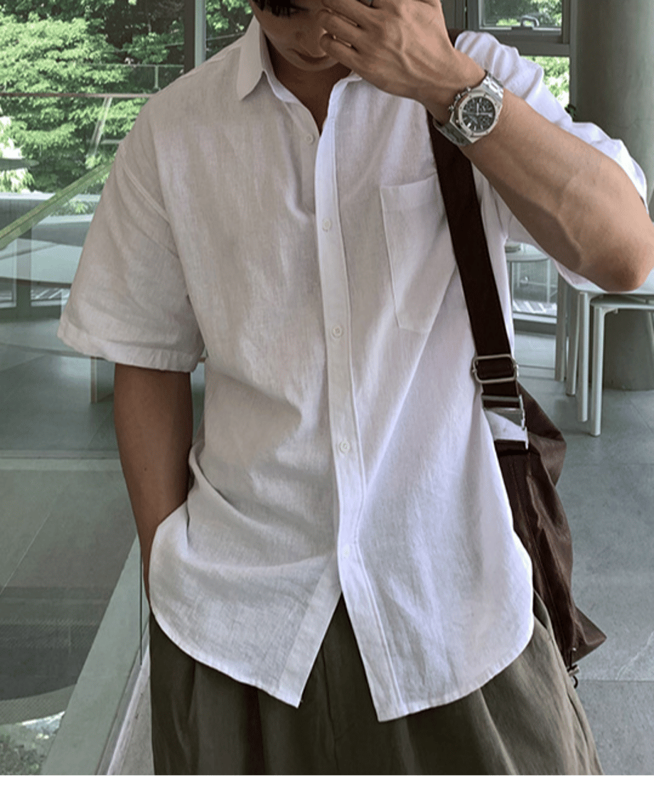 ホワイトのリネン半袖シャツのボタンを少し開け、腕時計をした左手を顔に添えて立つ男性の正面からのカット。
