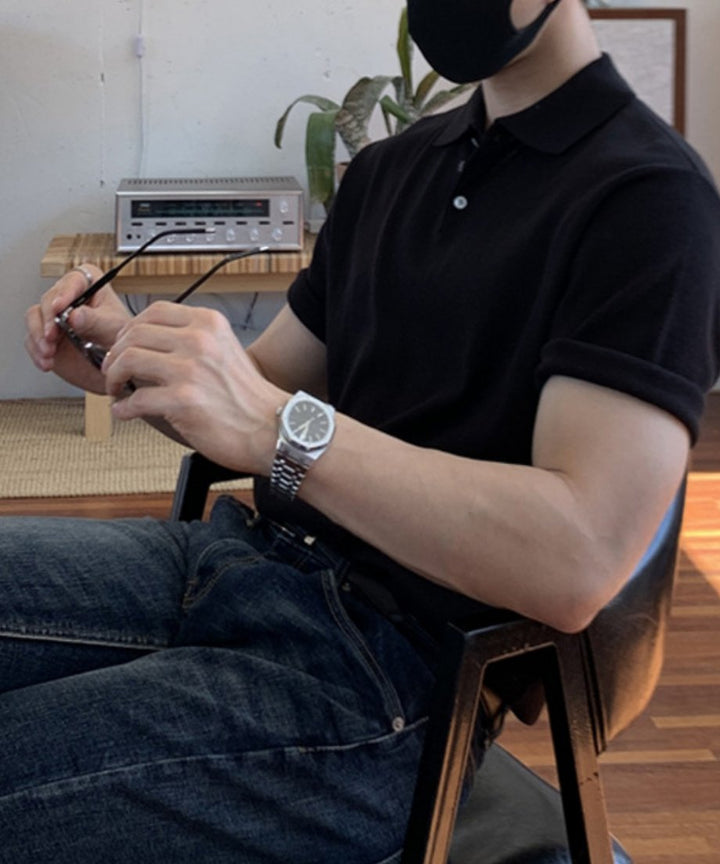 黒いマスクを着用し、ブラックの半袖ニットポロシャツにインディゴブルーのデニムを合わせ、眼鏡を手に持って椅子に座る男性のクローズアップ写真。