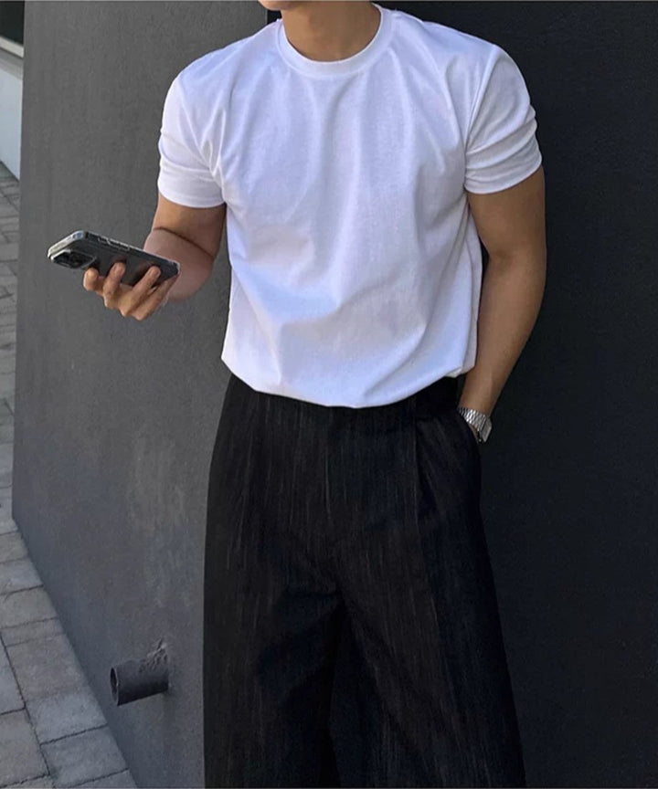 白い半袖クルーネックTシャツに黒いワイドスラックスを合わせ、スマートフォンを手にして壁に寄りかかる男性の斜め前からの立ち姿。