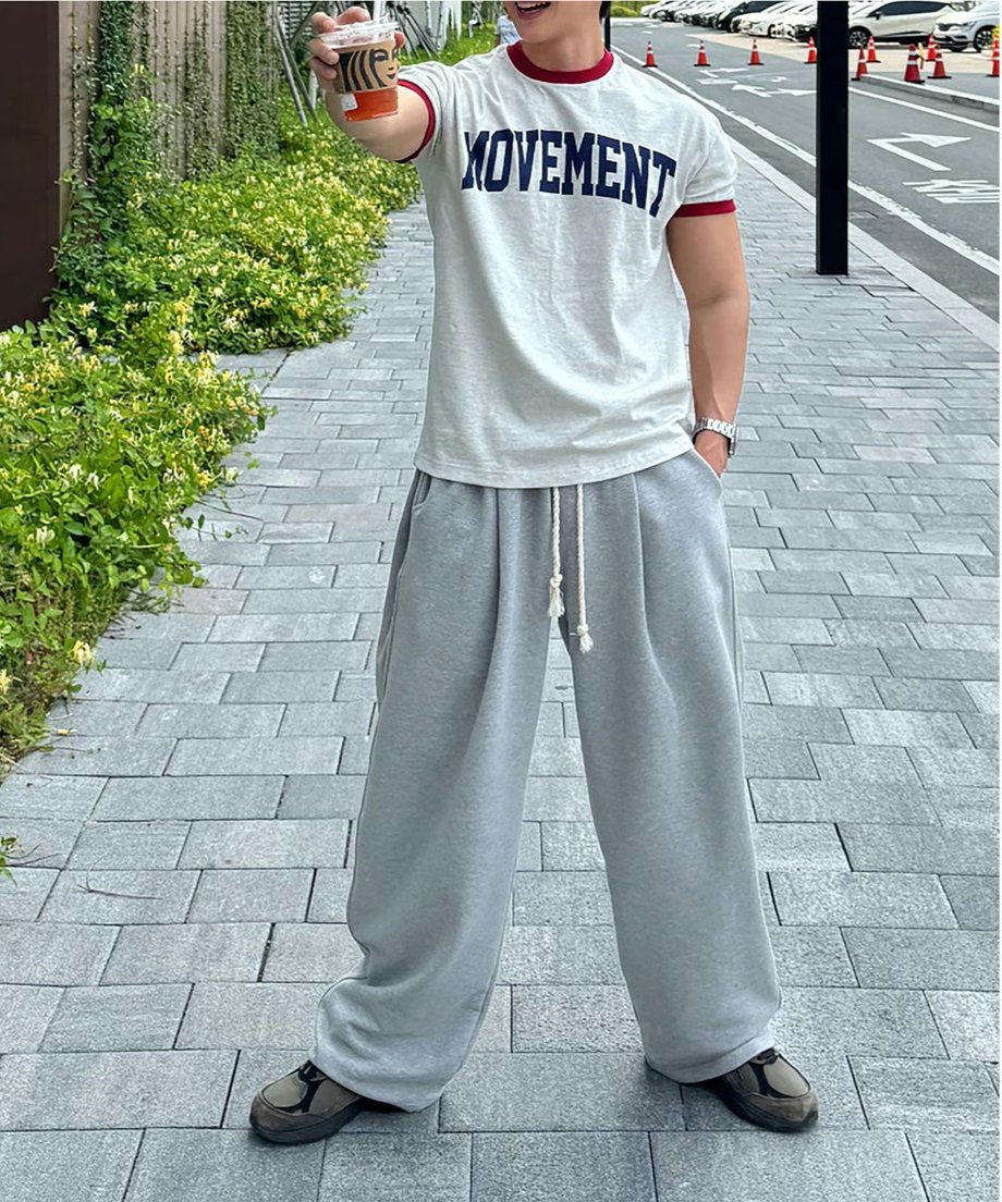 オートミール地のリンガーTシャツを着用し、屋外の石畳の上でドリンクを持ちながら、足を広げてポーズを取る男性の全身スタイリング写真。