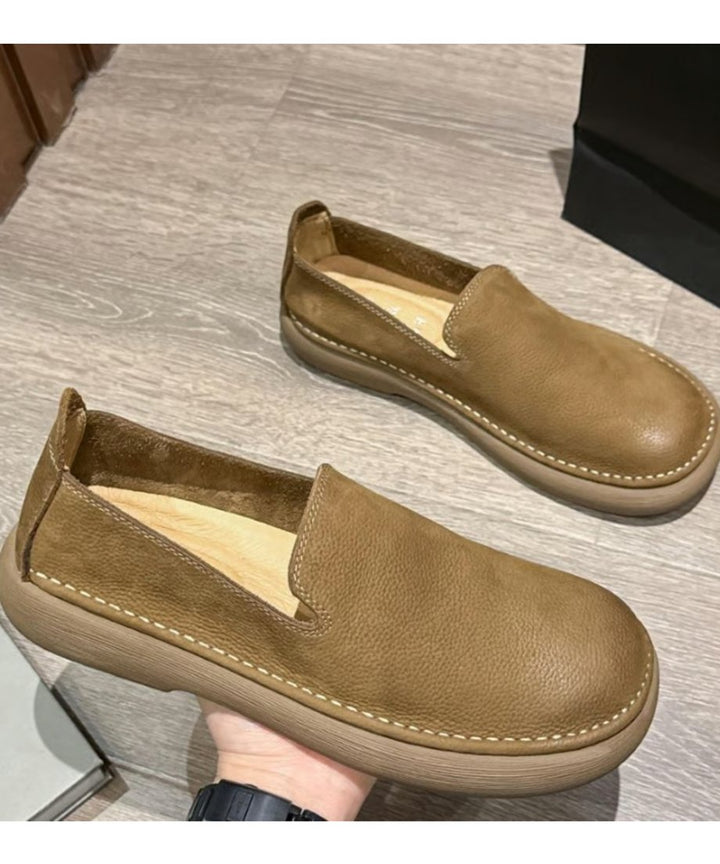 木目調の床の上で、シボ感のあるカーキのレザー調スリッポンシューズを手に持っているカット。