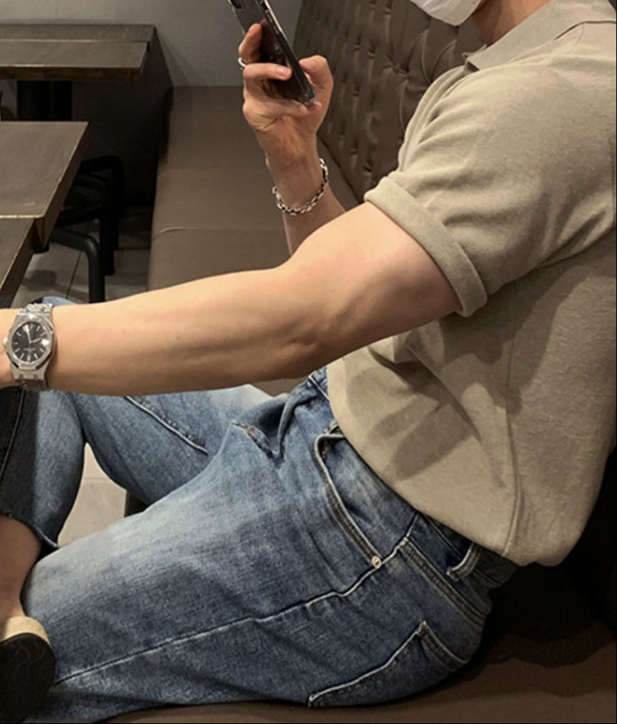 カフェのソファー席で、ウォームベージュの半袖ニットポロシャツにライトウォッシュデニムを合わせ、スマホを見つめる男性の横顔と腕のラインを捉えたカット。