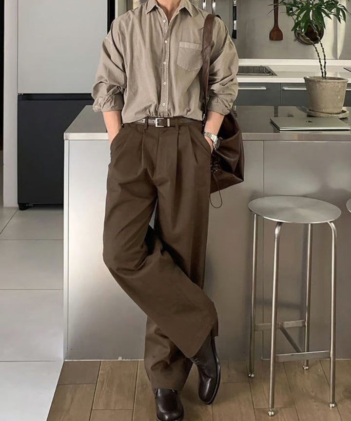 明るい室内のキッチンカウンター前で、ベージュの長袖シャツにブラウンのツータックワイドスラックスをタックインしてスタイリッシュに立つ男性の全身カット。