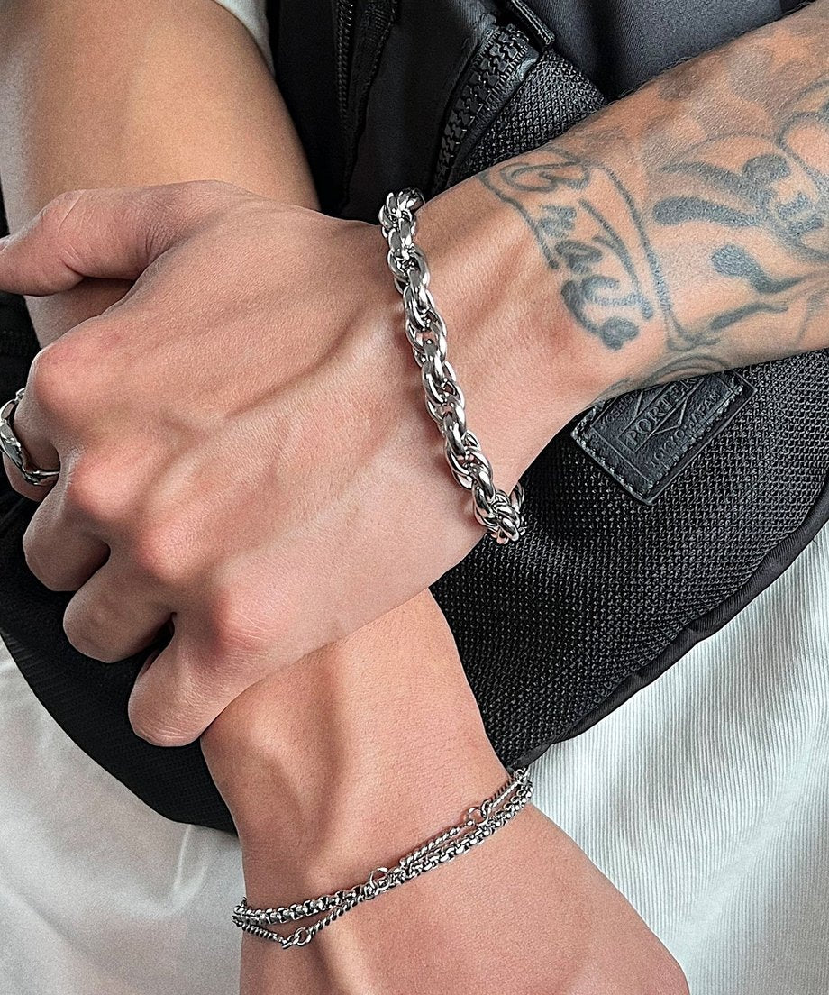 黒いバッグを背景に両腕を交差させ、ツイストチェーンブレスレットと細身の2連チェーンブレスレットの組み合わせによるこなれたレイヤードスタイルを提案するカット
