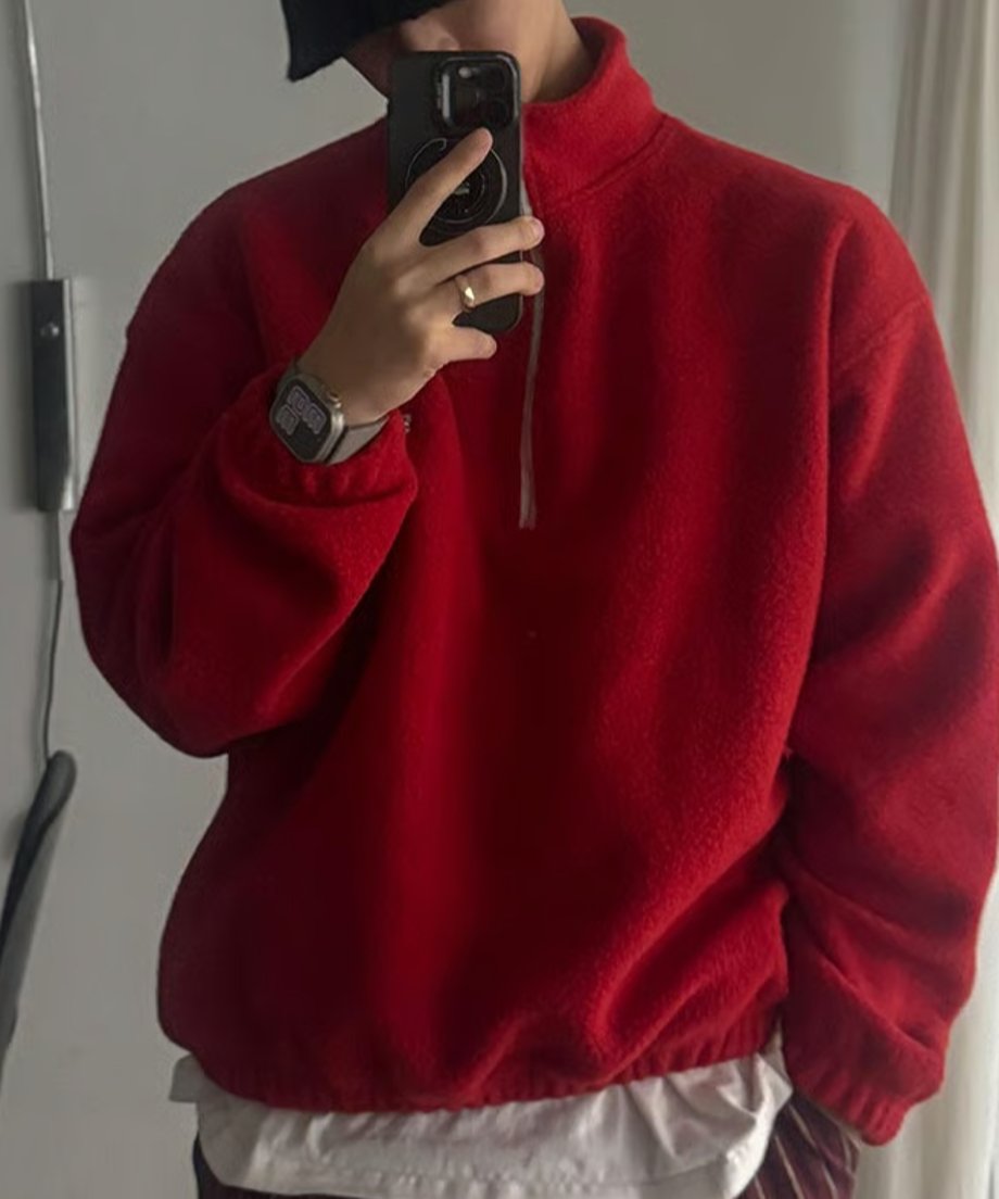 レッドのハーフジップフリースを着用した男性が、スマートフォンを鏡にかざして自撮り（セルフィー）をしている上半身カット