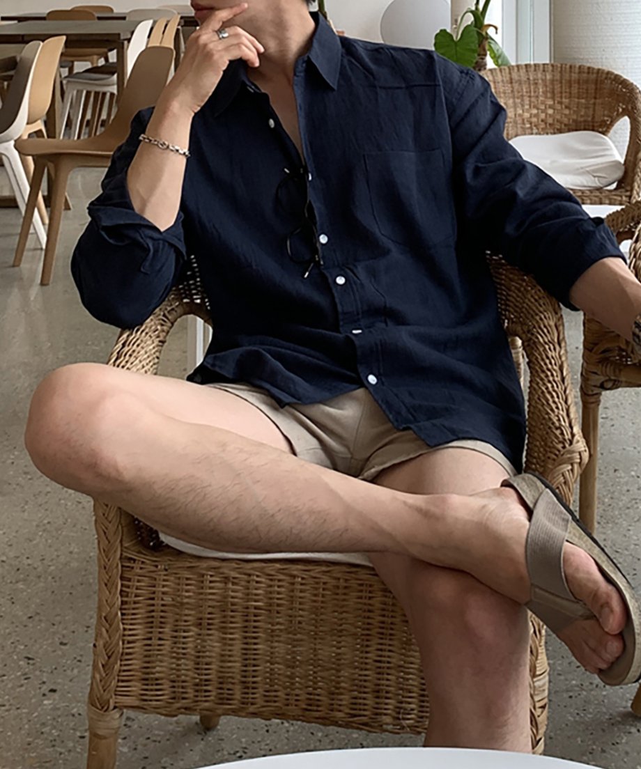 ネイビーブルーの長袖リネンシャツの袖をまくり、ラタンチェアで足を組んでリラックスする男性のポーズ写真。