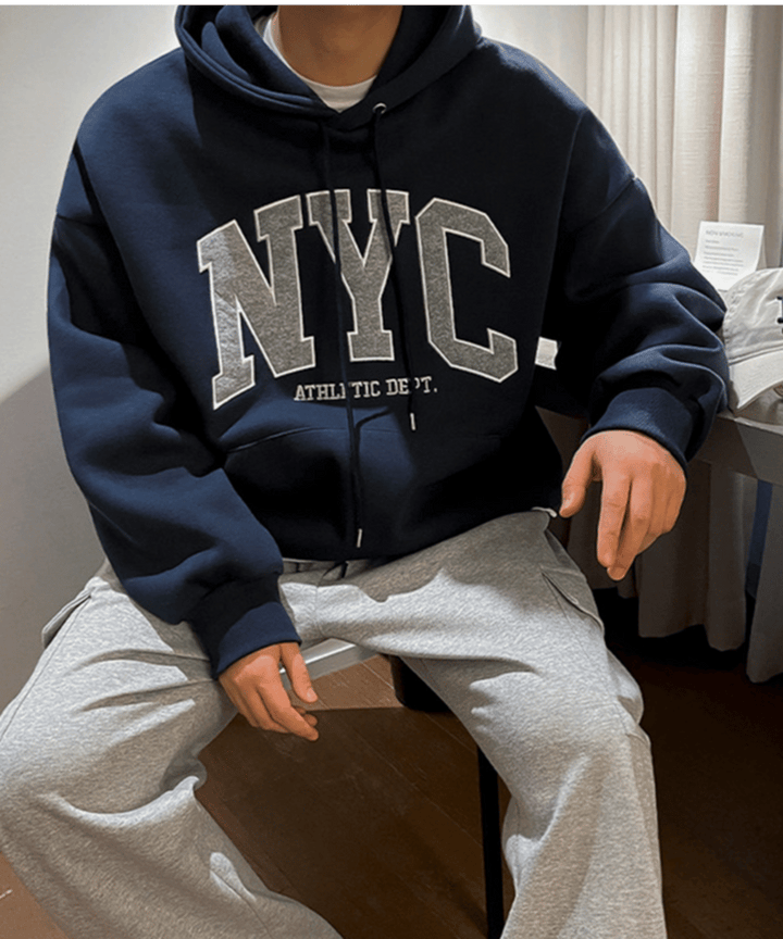 ネイビーブルーのロゴパーカーとグレーのカーゴパンツを着用し、椅子に腰掛けてリラックスする男性のスタイリッシュなスナップカット。