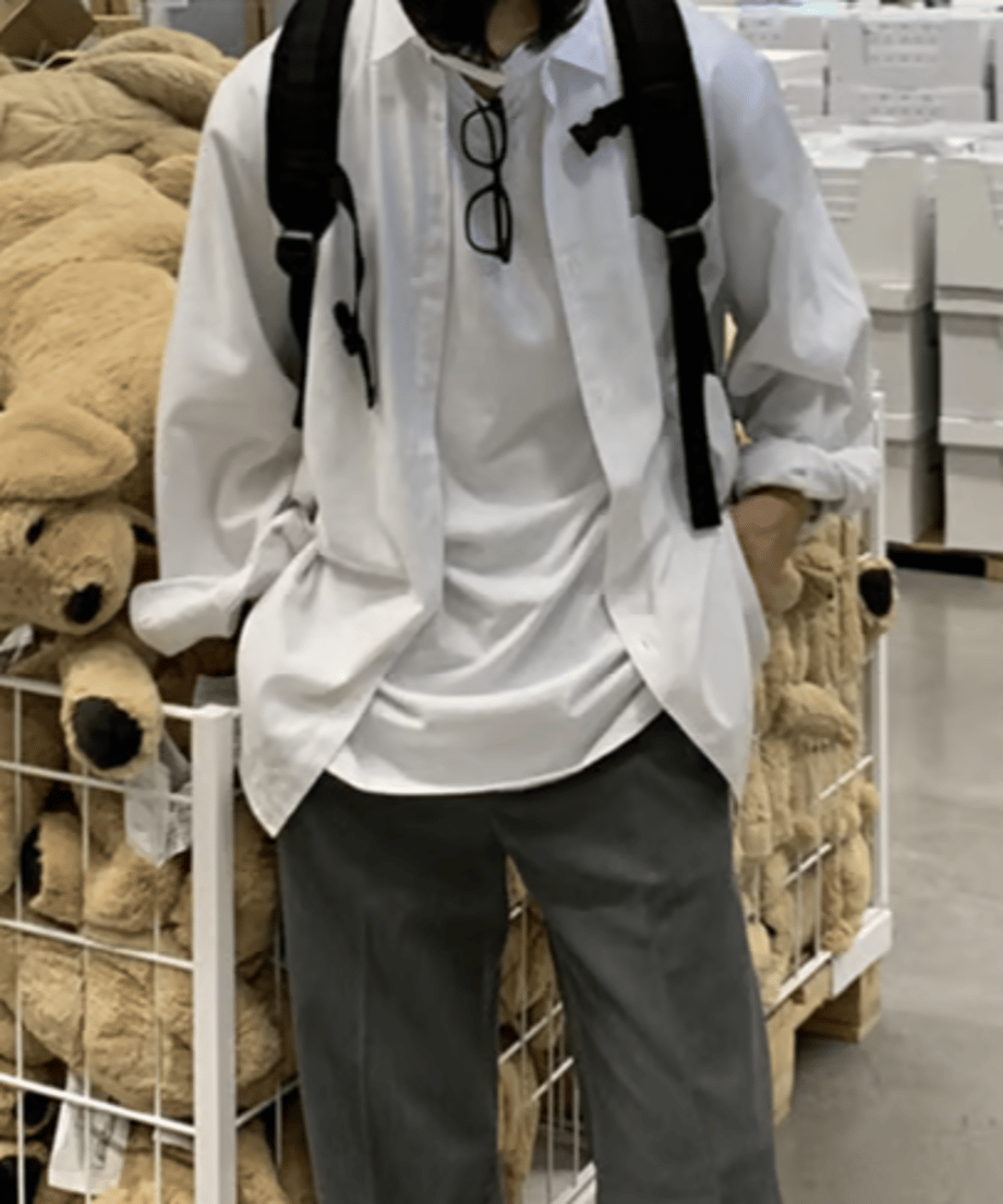 白い長袖シャツをTシャツの上に羽織り、グレーのパンツのポケットに両手を入れて、犬のぬいぐるみが並ぶカゴの前に立つ男性の正面スタイリング。