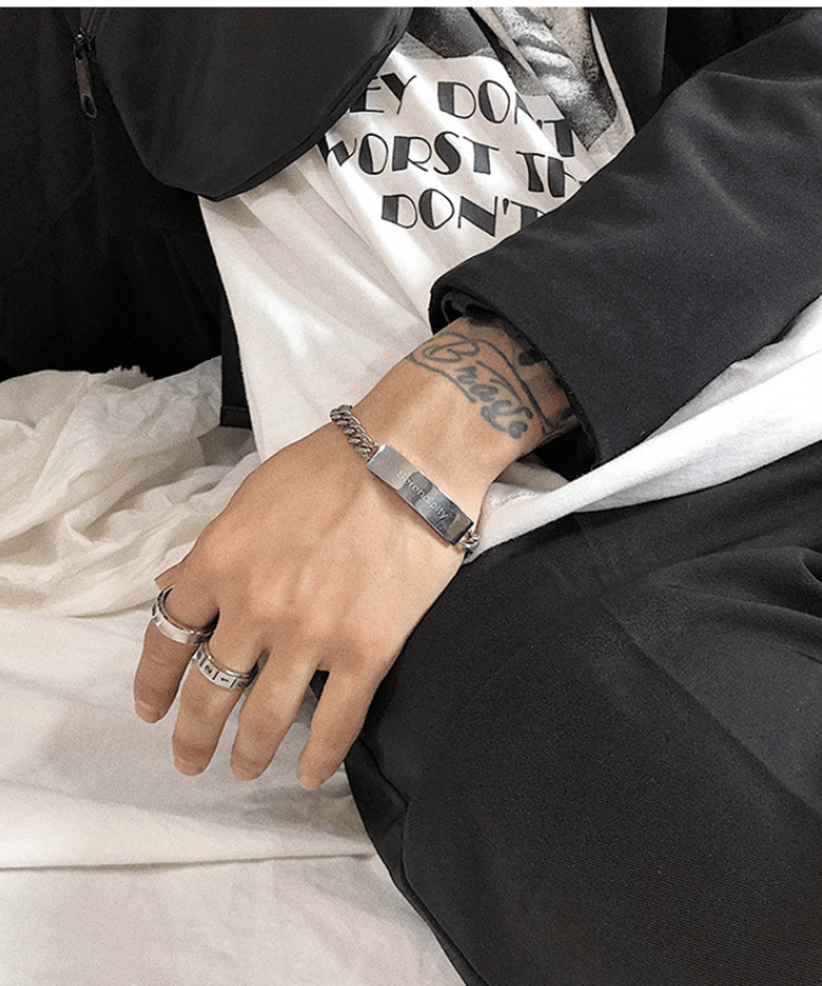 黒いスラックスを穿いて座る男性の手元を捉えた、モノトーンコーデにシルバーのIDプレートブレスレットがシャープな輝きを添えるカット
