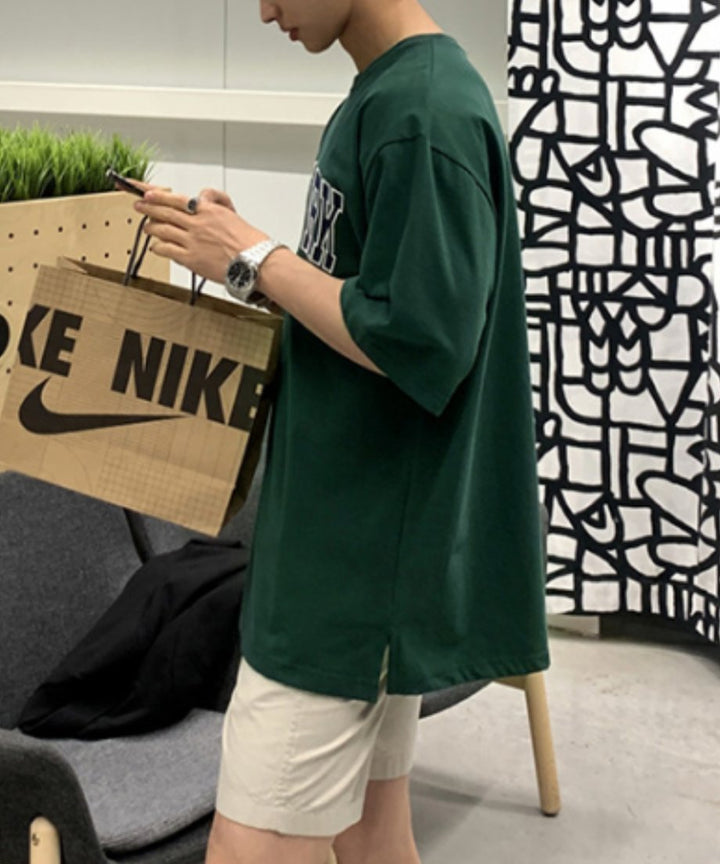 室内でダークグリーンのロゴTシャツとベージュのハーフパンツを着用し、紙袋を持ってスマートフォンを操作する男性の横顔