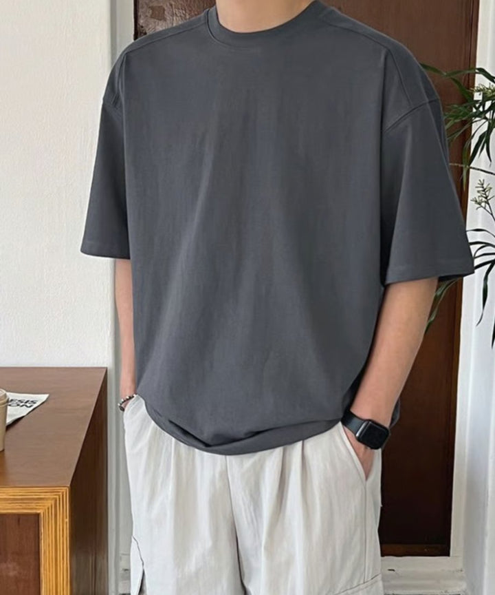 アイアングレーのクルーネックTシャツに白のカーゴパンツ、黒の腕時計を合わせ、ポケットに手を入れて佇む男性のチェストアップ。