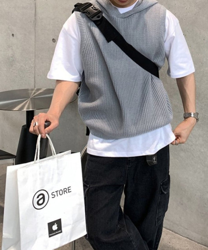 白Tシャツとグレーのフーディニットベストをレイヤードし、黒のカーゴパンツを穿いて白いショップバッグを手にする男性の立ち姿