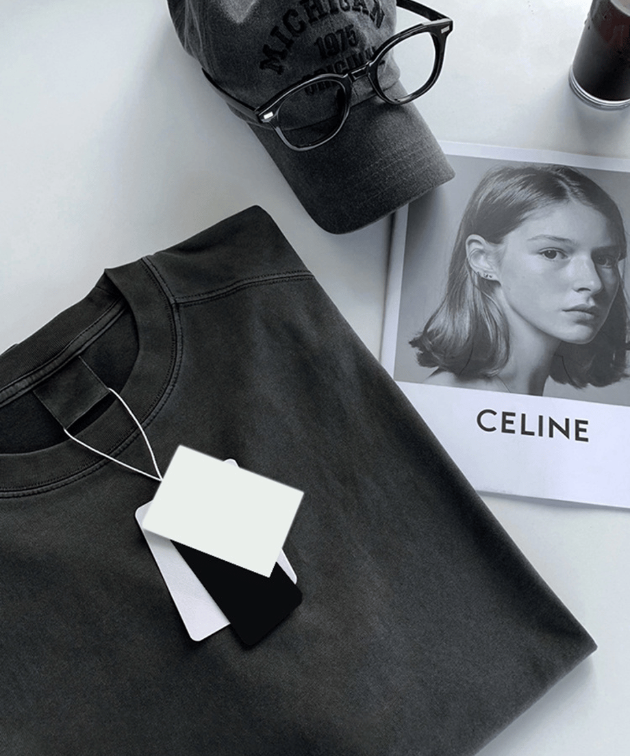 きれいに折りたたまれたアイアングレーの長袖Tシャツを中心に、モノトーンのブランド雑誌（CELINE）、チャコールグレーのロゴキャップ（MICHIGAN 1975）、黒縁のメガネを配置した、スタイリッシュなフラットレイ（置き画）カット。