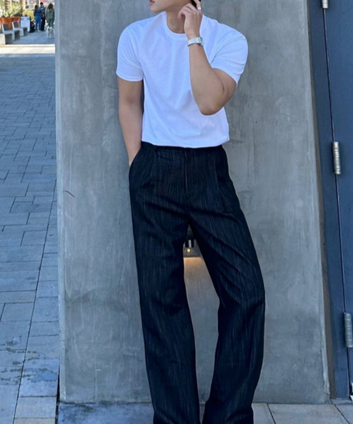 白い半袖クルーネックTシャツと黒いスラックスを着用し、屋外のコンクリート壁の前でポーズをとる男性の全身ショット。