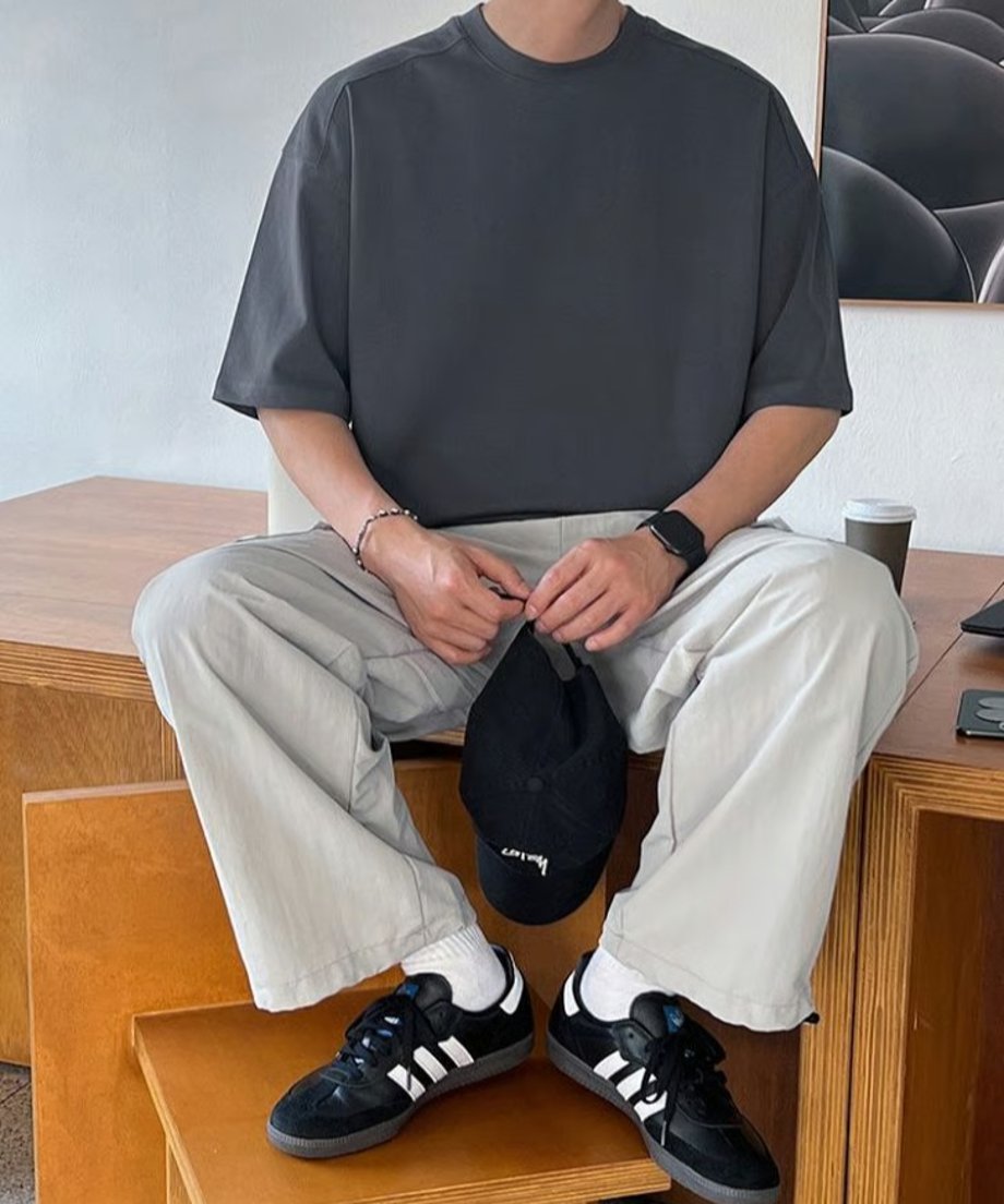 アイアングレーのオーバーサイズTシャツに白のカーゴパンツ、黒のスニーカーを合わせ、黒のキャップを両手で持ちながら木製の階段に腰掛ける男性の正面全身スタイリング。