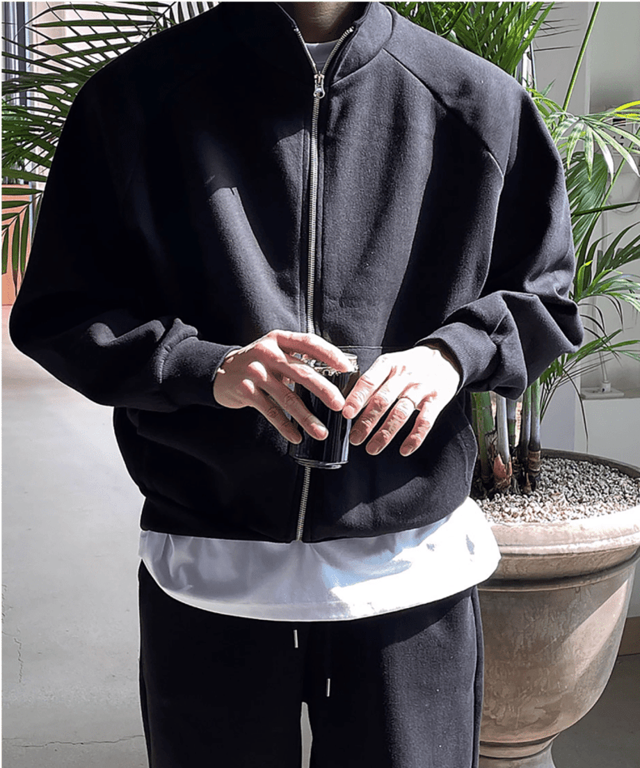 光が差し込む場所で、ブラックのジップアップジャケットのセットアップを着用し、インナーの白Tシャツを覗かせている男性の正面アップ