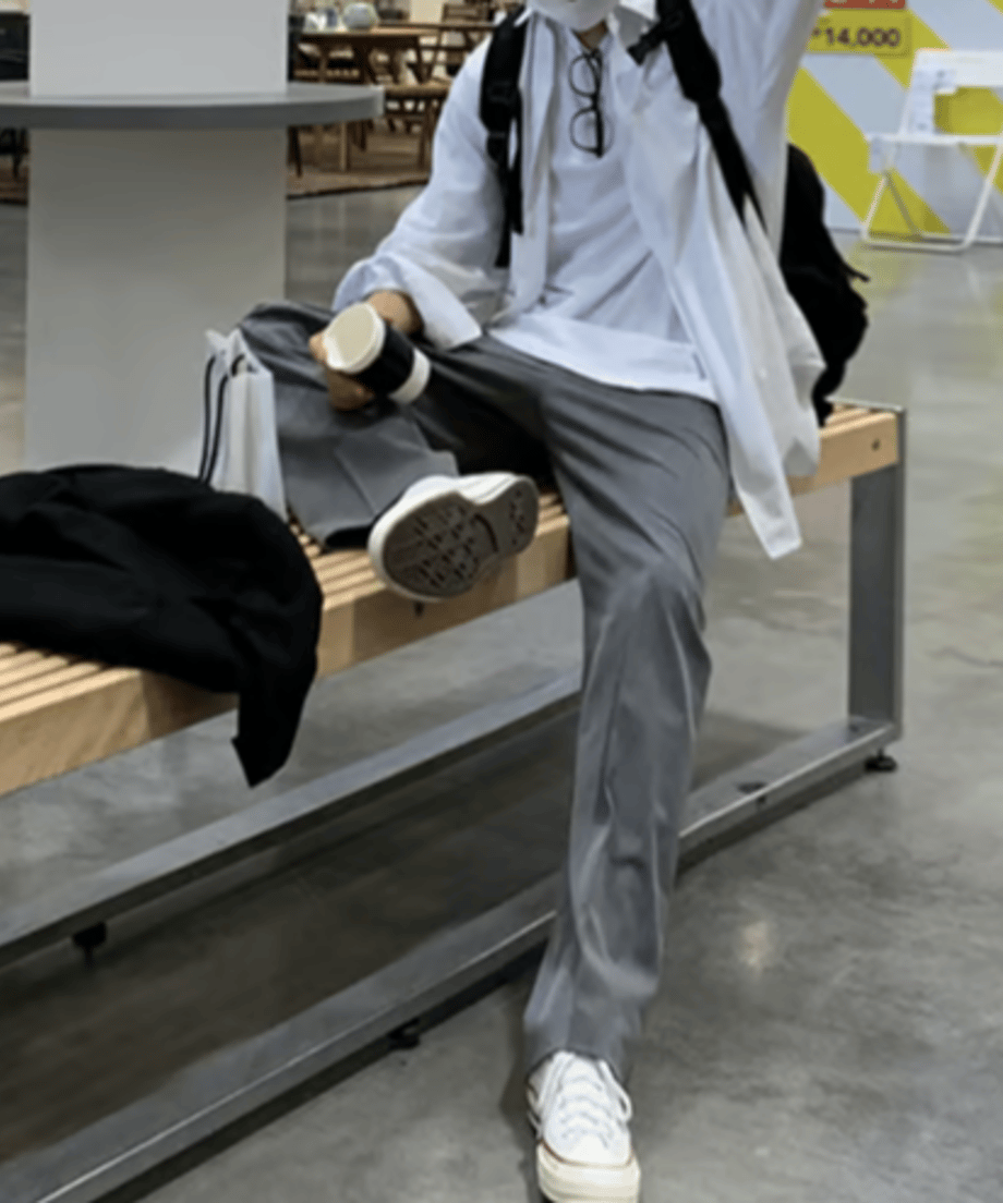白い長袖シャツにグレーのワイドパンツ、白いスニーカーを合わせ、ベンチに腰掛けてドリンクを持ちながら足を組む男性の全身スタイリング。
