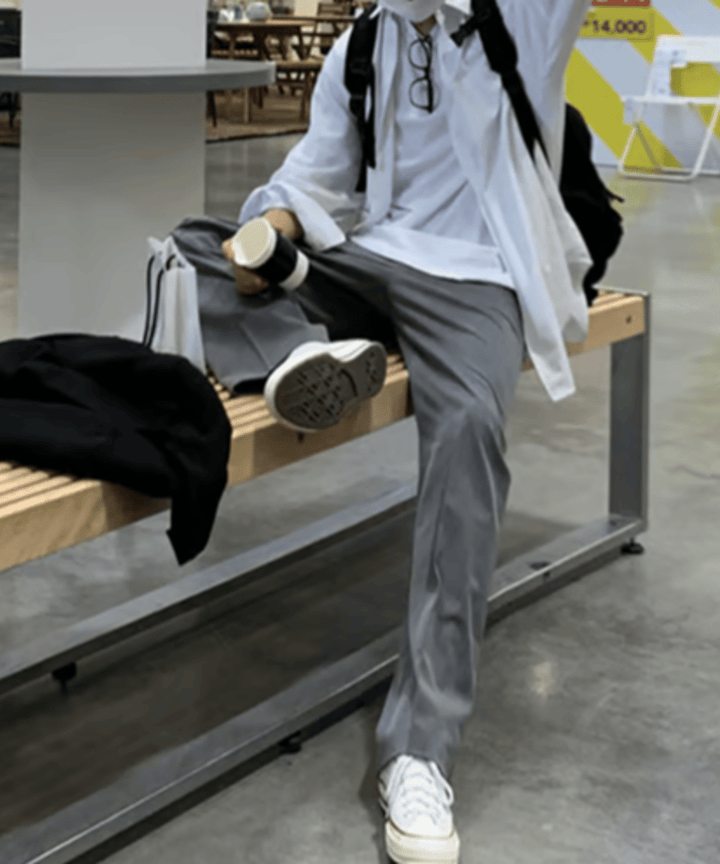 白い長袖シャツにグレーのワイドパンツ、白いスニーカーを合わせ、ベンチに腰掛けてドリンクを持ちながら足を組む男性の全身スタイリング。