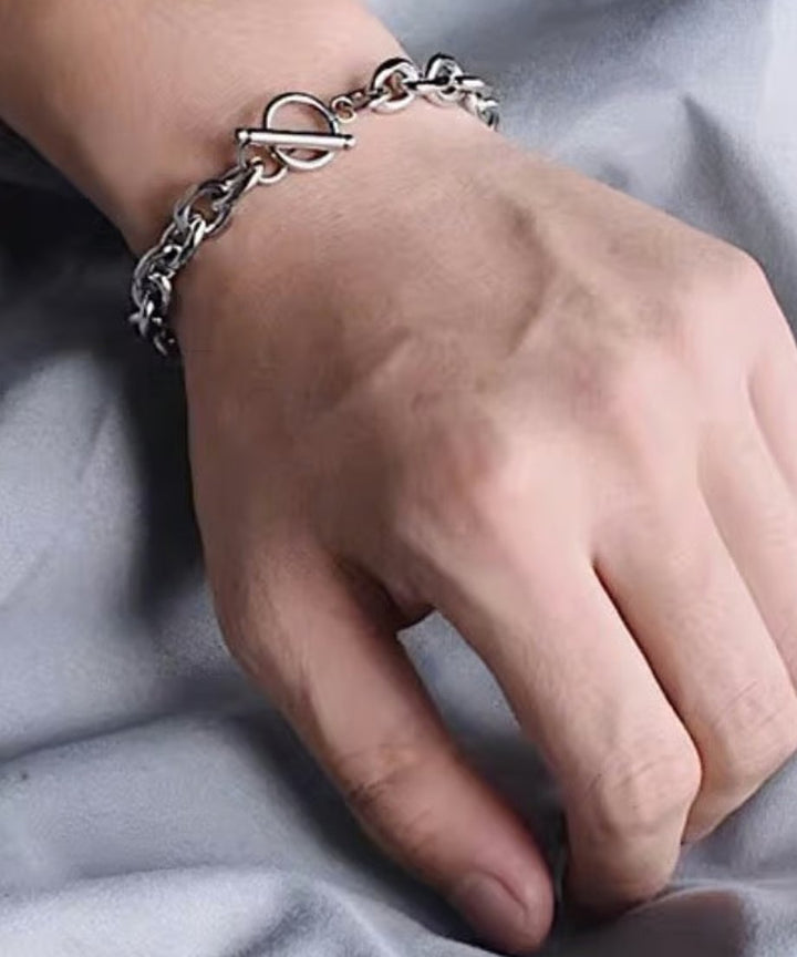 グレーの背景をバックに、男性の手首に程よいゆとりを持って巻き付いたシルバーチェーンとマンテルパーツの立体感を捉えたクローズアップ。