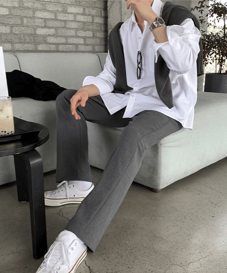 ホワイトのシャツにグレーのパンツ、白いスニーカーを合わせた男性がソファに腰掛け、脚を伸ばしているローアングルからのサイド全身スタイリング。