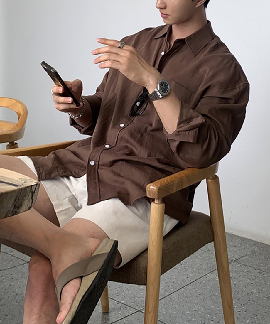 木製のアームチェアに座り、ブラウンの長袖リネンシャツを着てスマートフォンを見つめる男性のポートレート写真。