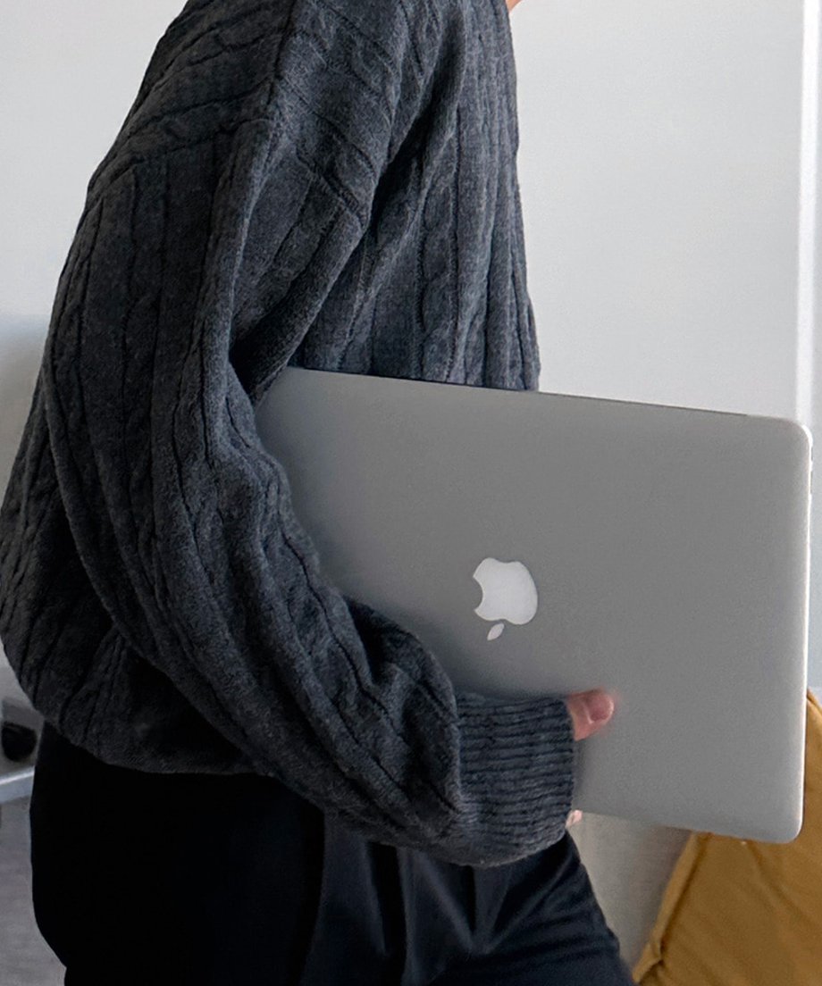 ダークグレーのニットを着用し、シルバーのノートパソコン（MacBook）を片手に抱えて歩く男性のスタイリッシュな手元アップカット。