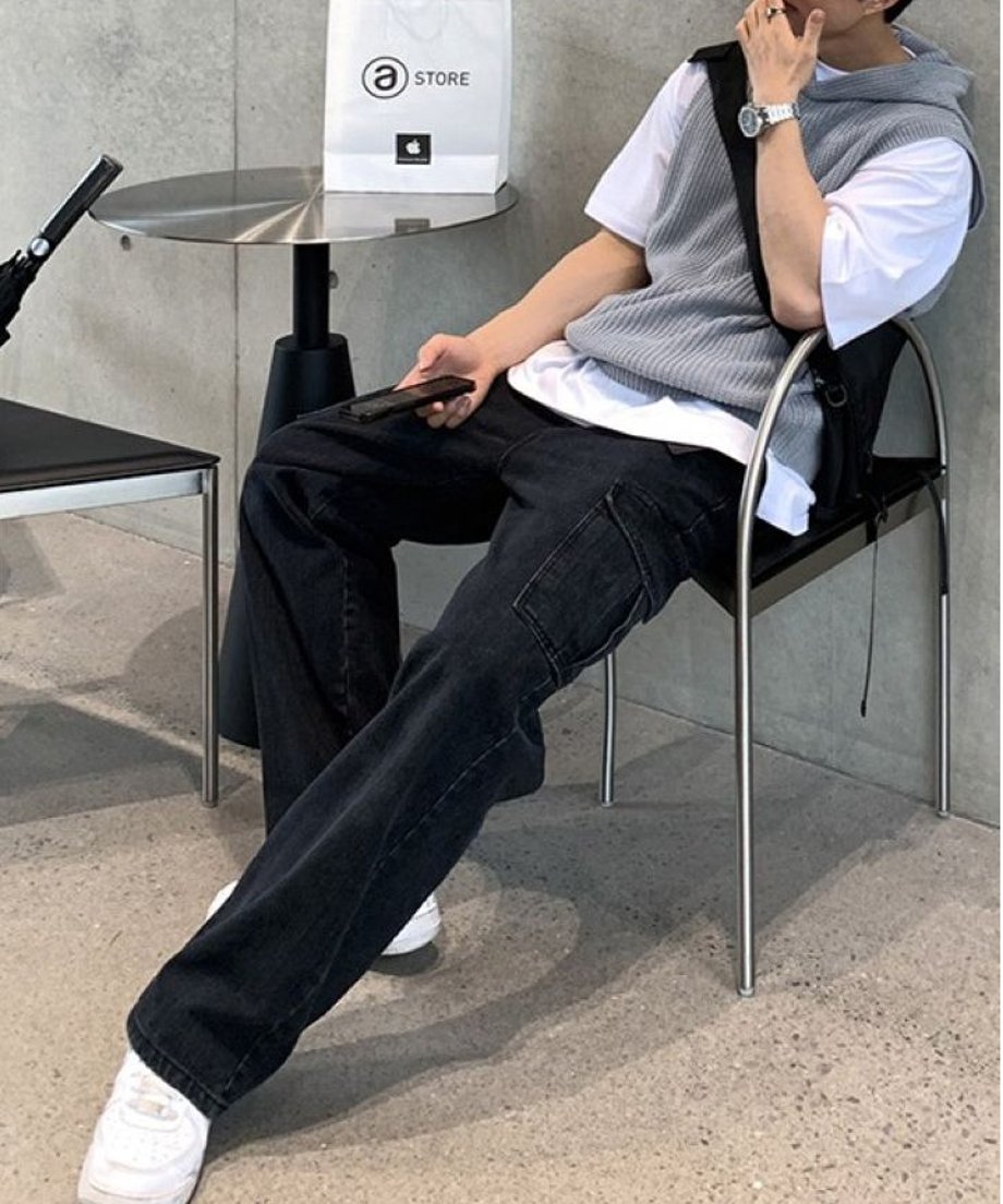 室内の椅子に腰掛け、白Tシャツにグレーのフード付きベスト、黒のワイドパンツを合わせたリラックスした佇まいの男性