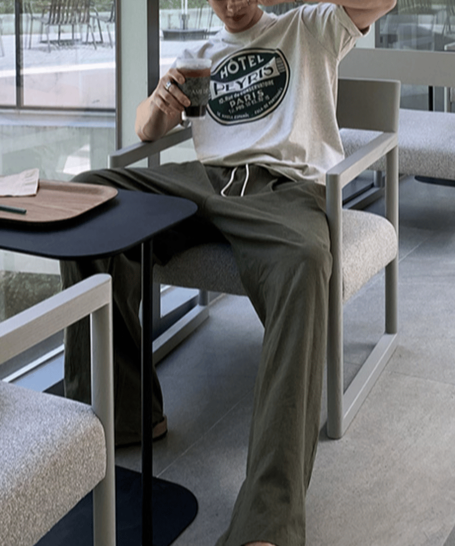 グレーのサークルロゴTシャツとオリーブグリーンのイージーパンツを着用した男性が、カフェのテラス席風の椅子にリラックスして腰掛け、アイスコーヒーを手にしているカット。