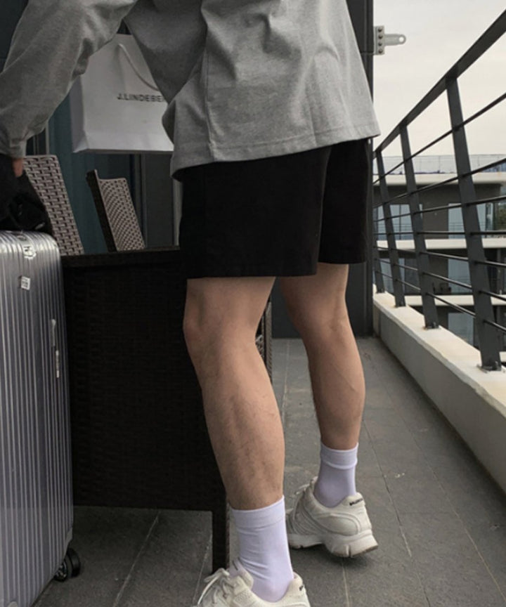 テラスのバルコニーで、グレーの長袖トップスに黒のショートパンツ、白ソックスと白スニーカーを合わせ、シルバーのスーツケースの横に立つ男性のバックサイドアングル
