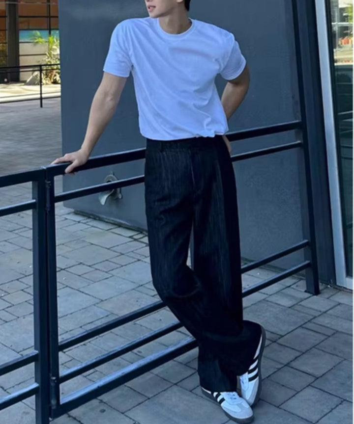 白い半袖クルーネックTシャツに黒いパンツ、足元に白いスニーカーを合わせ、黒い手すりに寄りかかる男性の全身ショット。