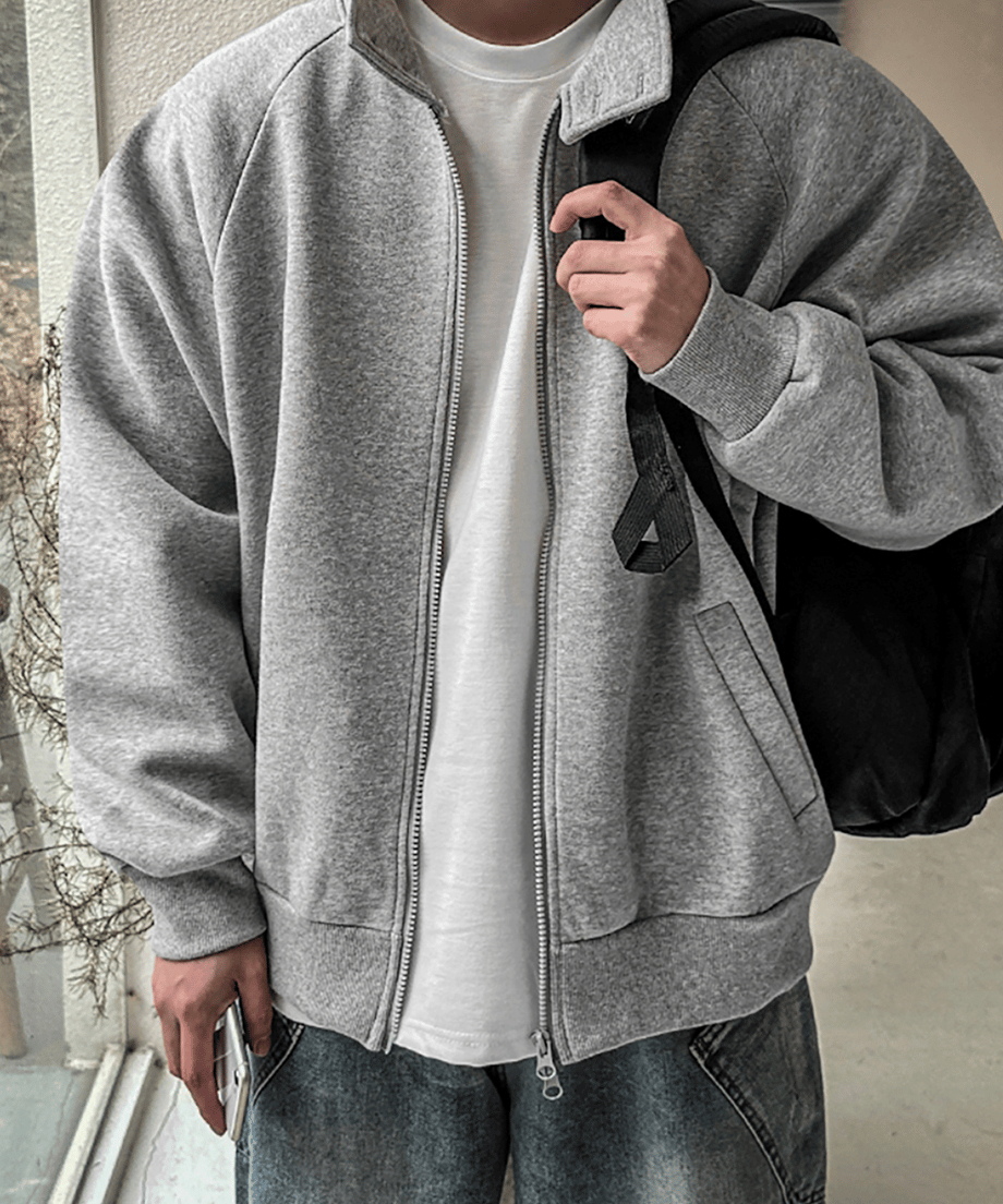 グレーのジップアップジャケットのジッパーを少し開け、インナーの白Tシャツを見せてリュックサックのストラップを掴む男性のアップ