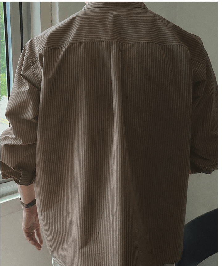 ブラウンのストライプ長袖シャツを着用し、窓際に立つ男性を後ろから捉えたバックシルエット。