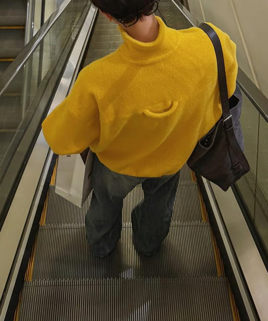 マスタードイエローのハーフジップフリースを着用した男性を、エスカレーターの上から見下ろすように撮影したバックカット。