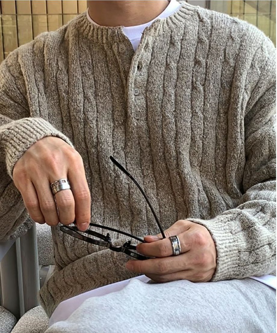 ベージュのヘンリーネックニットの首元のボタンディテールや立体的なケーブル編みの質感、手元のシルバーリングを近くで捉えたアップカット。
