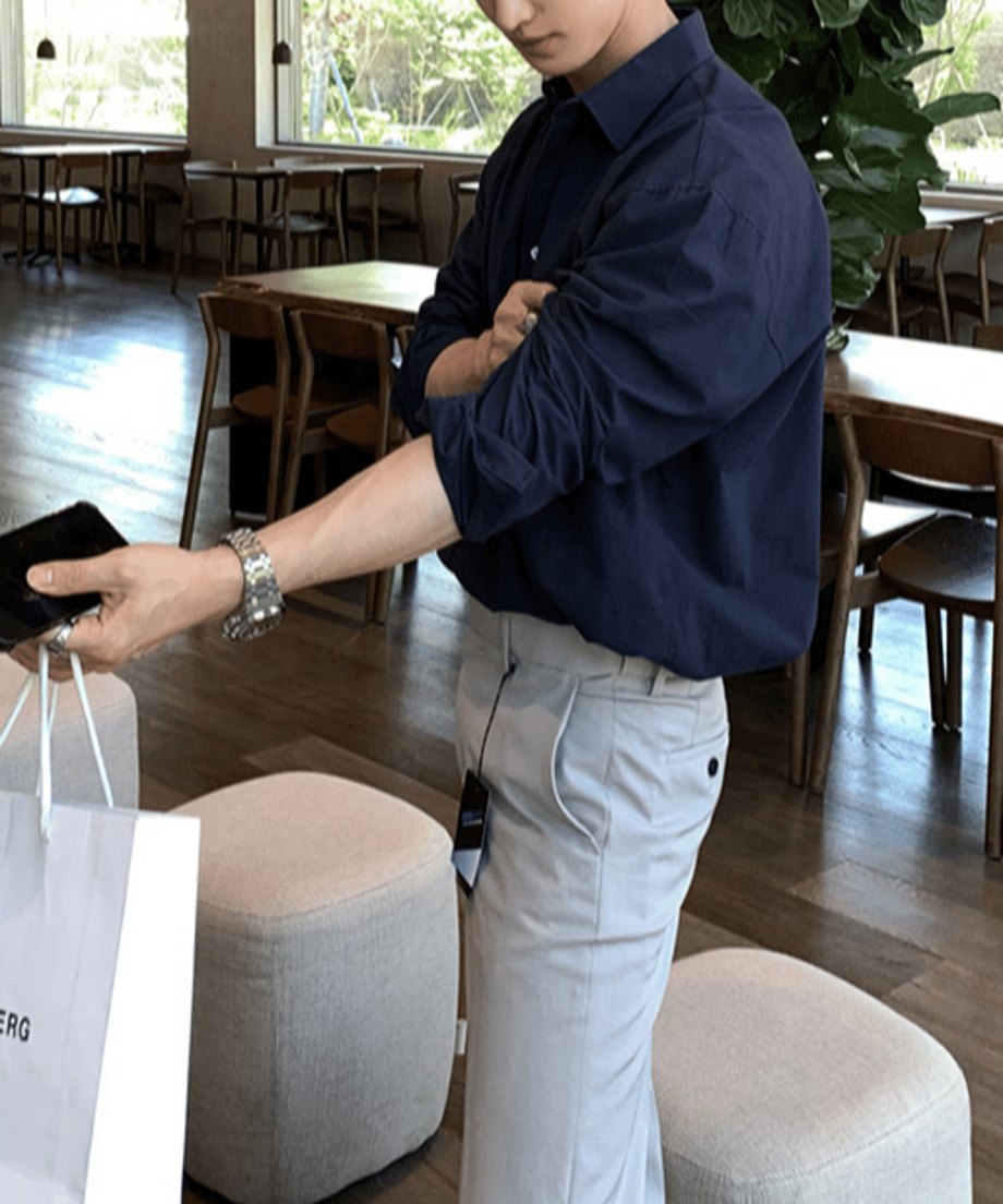カフェの店内で、白いショップバッグを手に持ちながらネイビーシャツの袖をまくり上げている男性のサイドカット。