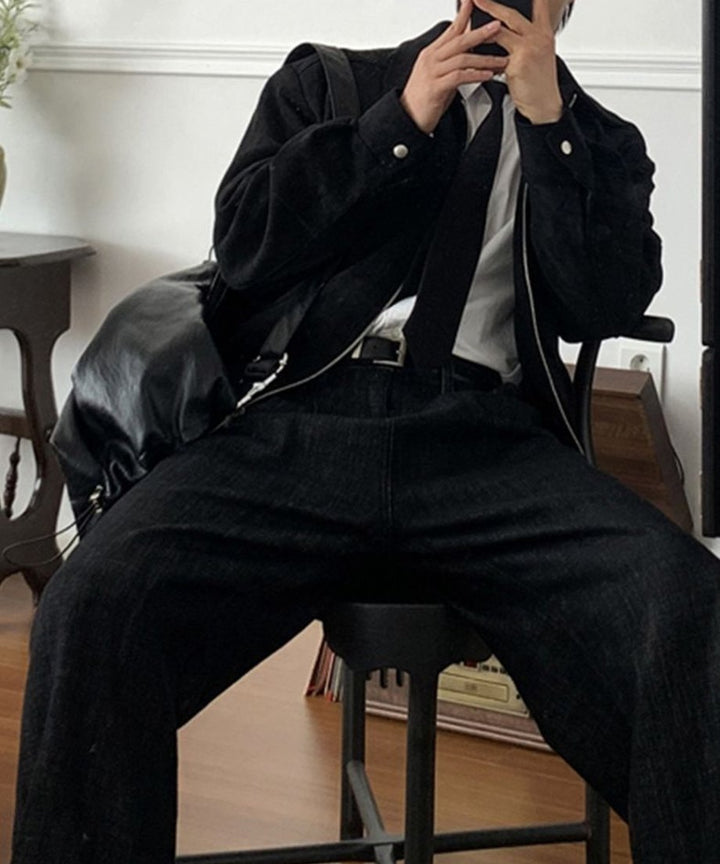 室内のスツールに深く腰掛け、ブラックのスエード調ブルゾンとワイドパンツを着用してスマートフォンを顔の前に構える男性のスタイリング