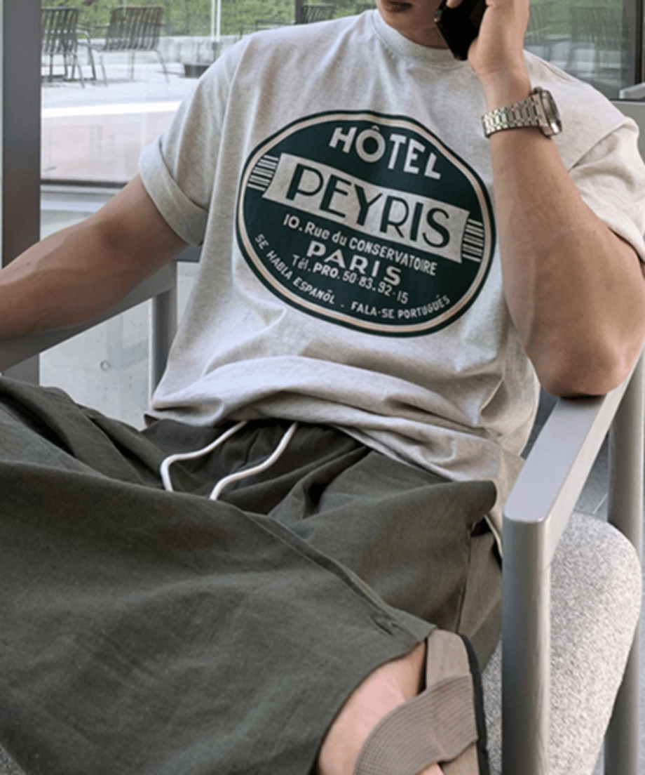 グレーのサークルロゴTシャツにオリーブグリーンのパンツを合わせ、椅子に座りながらスマートフォンで通話をしている男性のクローズアップカット。