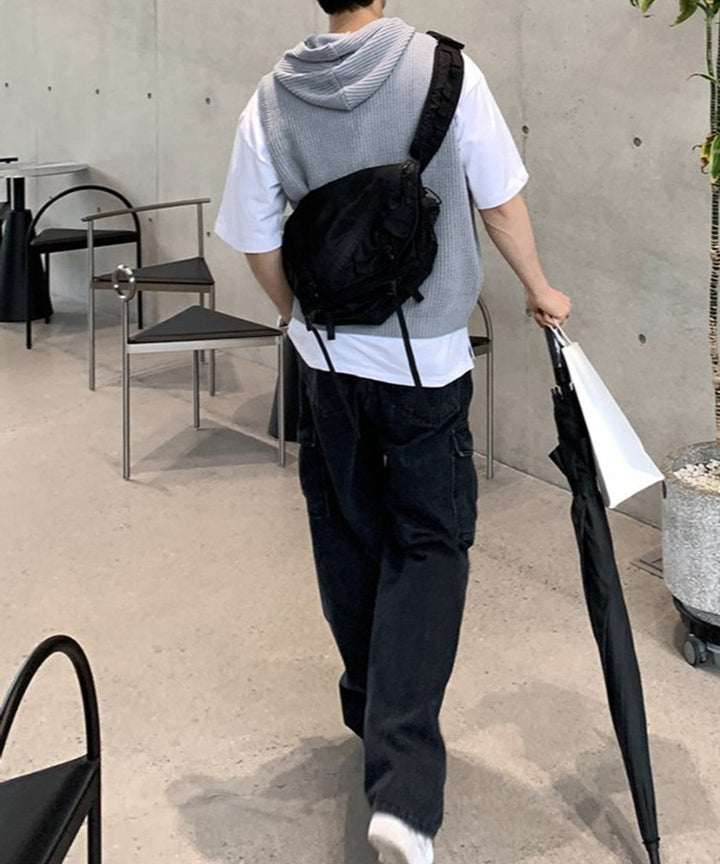 グレーのフード付きニットベストに黒のショルダーバッグを背負い、右手に黒い傘、左手にショップバッグを持って歩く男性の後ろ姿