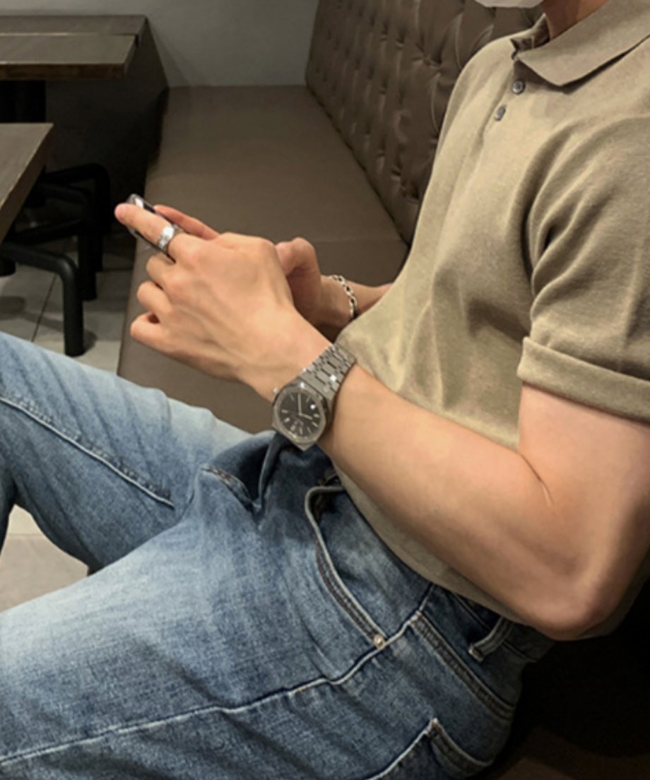 カフェのブラウンのソファーに座り、ウォームベージュのニットポロシャツの袖を少しロールアップしてスマホを操作する男性の斜め前からのカット。