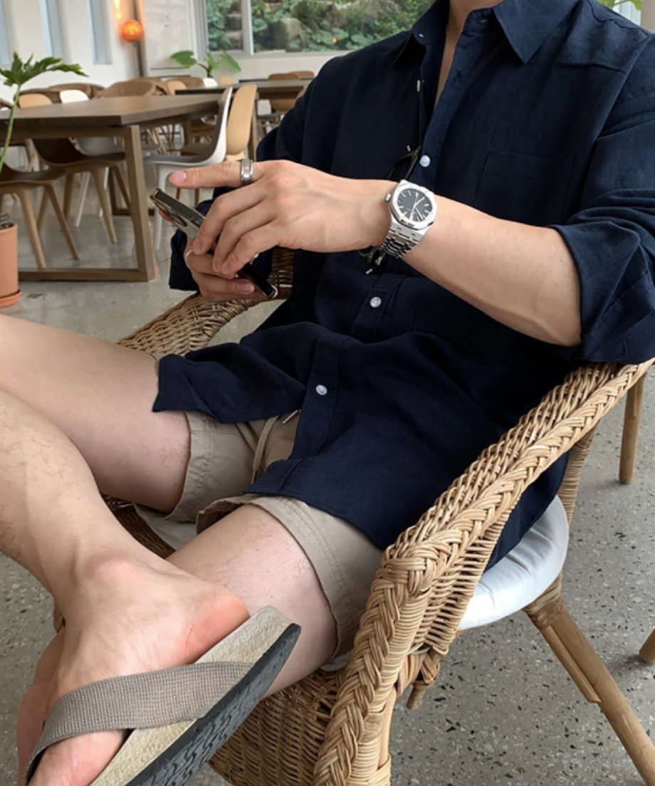 カフェのラタンチェアに座り、ネイビーブルーの長袖リネンシャツにベージュのショートパンツを合わせた男性がスマートフォンを操作している写真。