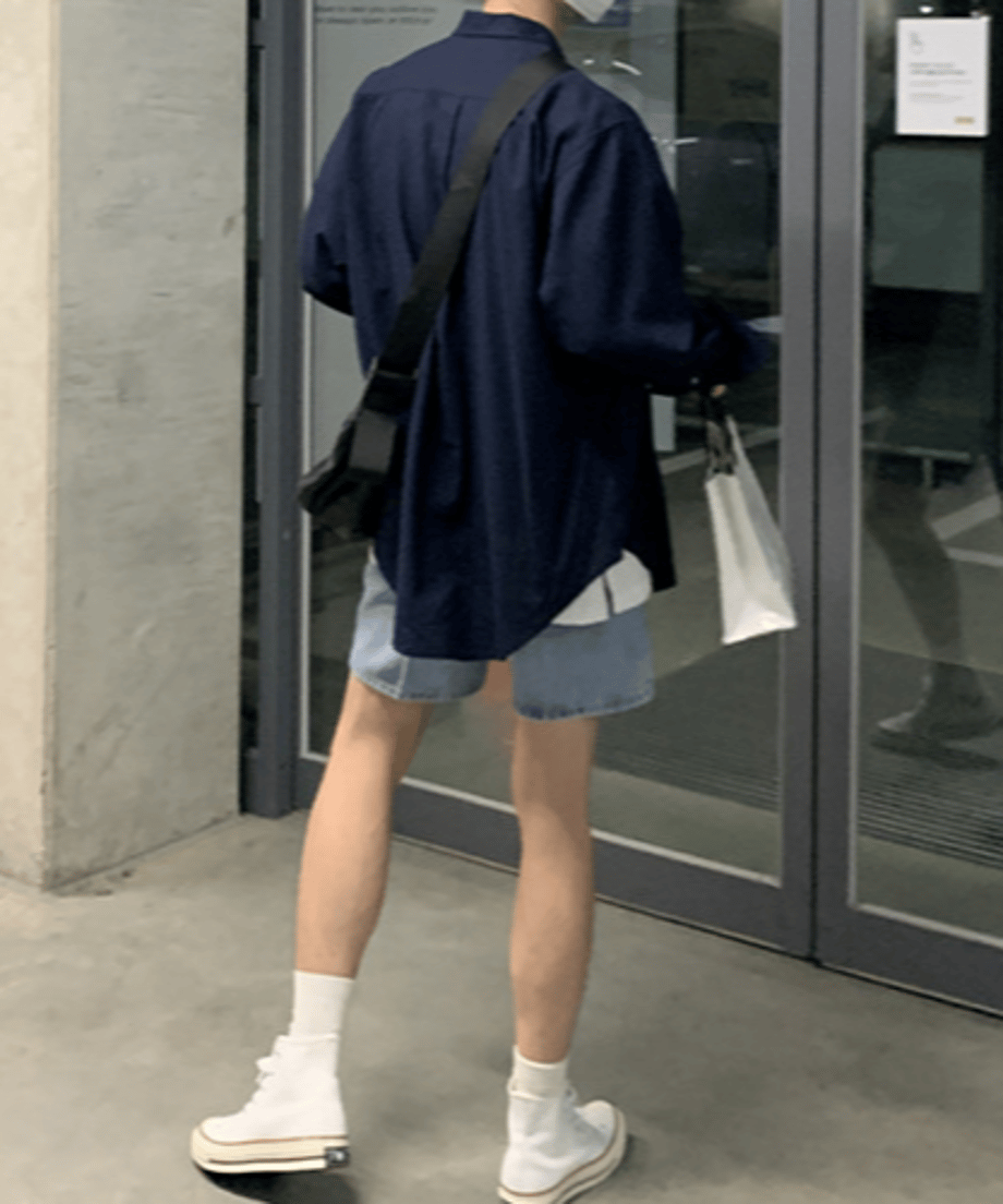 ネイビーのオーバーサイズ長袖シャツにデニムのショートパンツ、白いハイカットスニーカーを合わせ、白いショップバッグを持って歩く男性の後ろ姿。