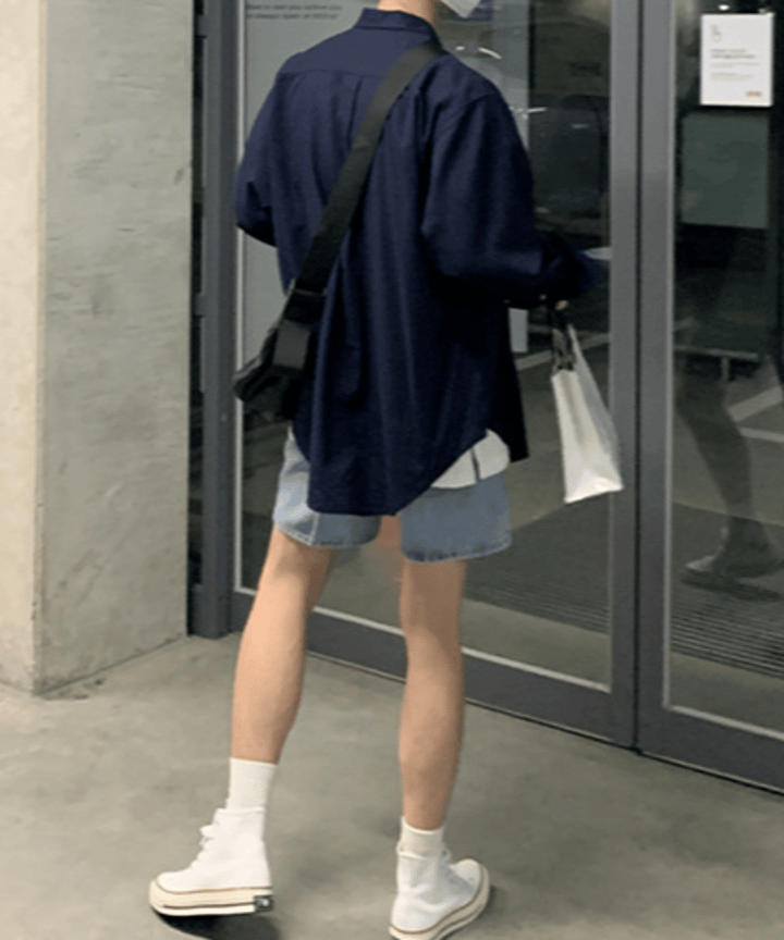 ネイビーのオーバーサイズ長袖シャツにデニムのショートパンツ、白いハイカットスニーカーを合わせ、白いショップバッグを持って歩く男性の後ろ姿。