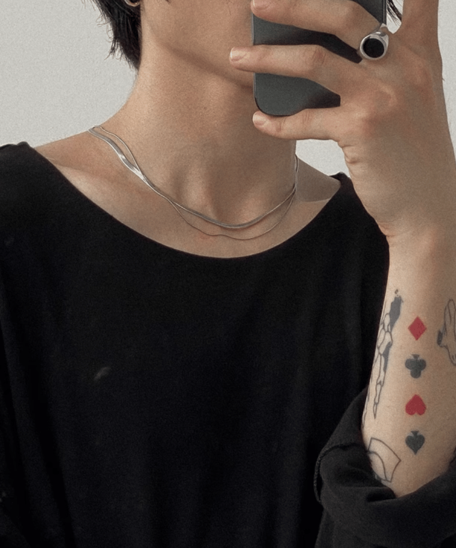 黒いトップスを着用した男性がスマホで自撮り（ミラーセルフィー）する手元と首元を捉えた、ストリートモードな着用イメージカット。